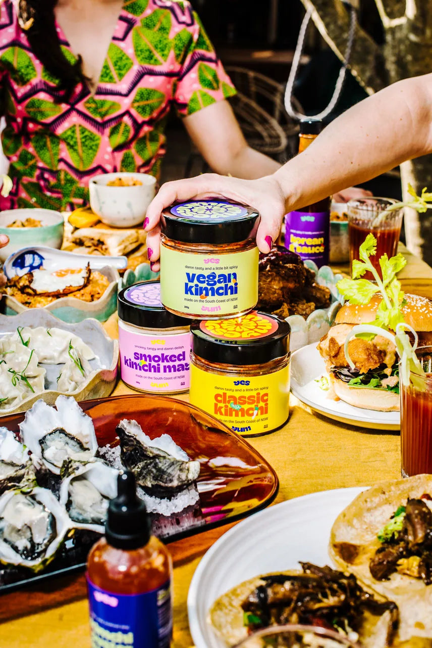Person reaching for a jar of vegan kimchi among various food items on a table.