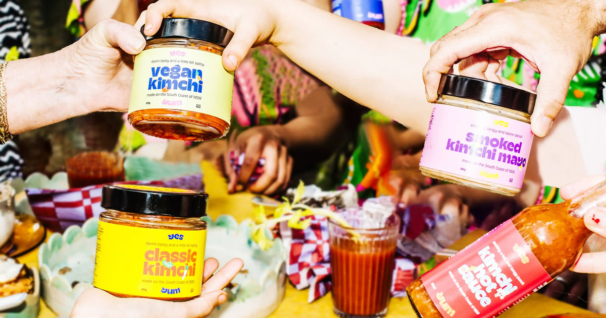 Three jars of different types of kimchi held by people at a table with food and drinks.