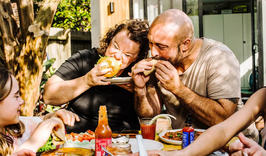 Two men eating sandwiches at a table with drinks and food around them.