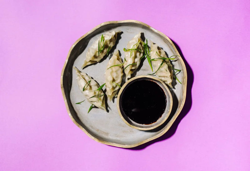 Dumplings with green herbs on a plate with a small bowl of dipping sauce against a pink background