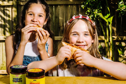 Two young girls eating pizza with jars of vegan and classic sauces on a table outdoors.