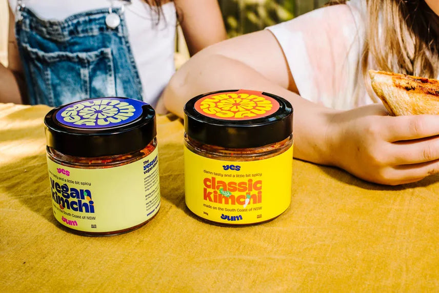 Two jars of vegan kimchi on a yellow surface with people in the background.