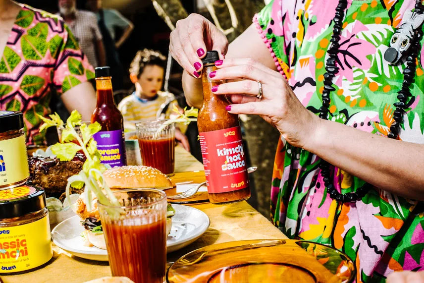 Person pouring hot sauce into a glass at a table with various condiments and drinks.