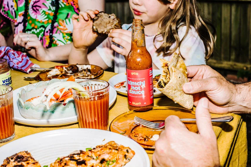 People enjoying a meal with pizza, drinks, and a bottle of kimchi hot sauce on a table.