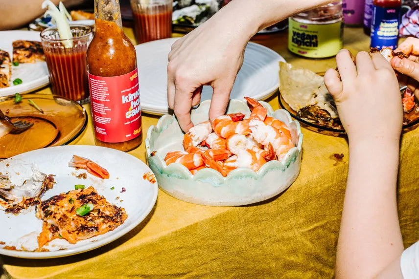 Person adding shrimp to a dish on a table with various items including a bottle of hot sauce.