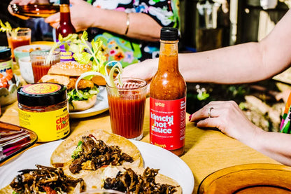 Table with food, drinks, and a bottle of 'YesYum Hot Sauce' in a casual setting.