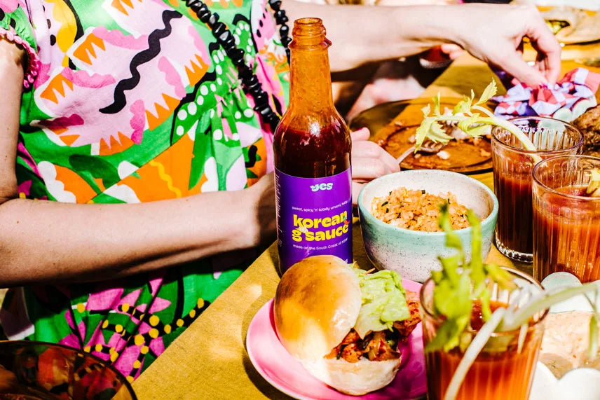 Person in colorful outfit with 'Korean & Sauce' bottle, food, and drinks on a table.