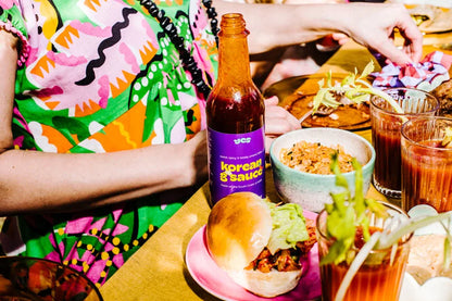 Person in colorful outfit with 'Korean & Sauce' bottle, food, and drinks on a table.