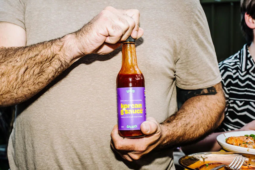 Person holding a bottle of sauce with a purple label, sitting at a table with food.