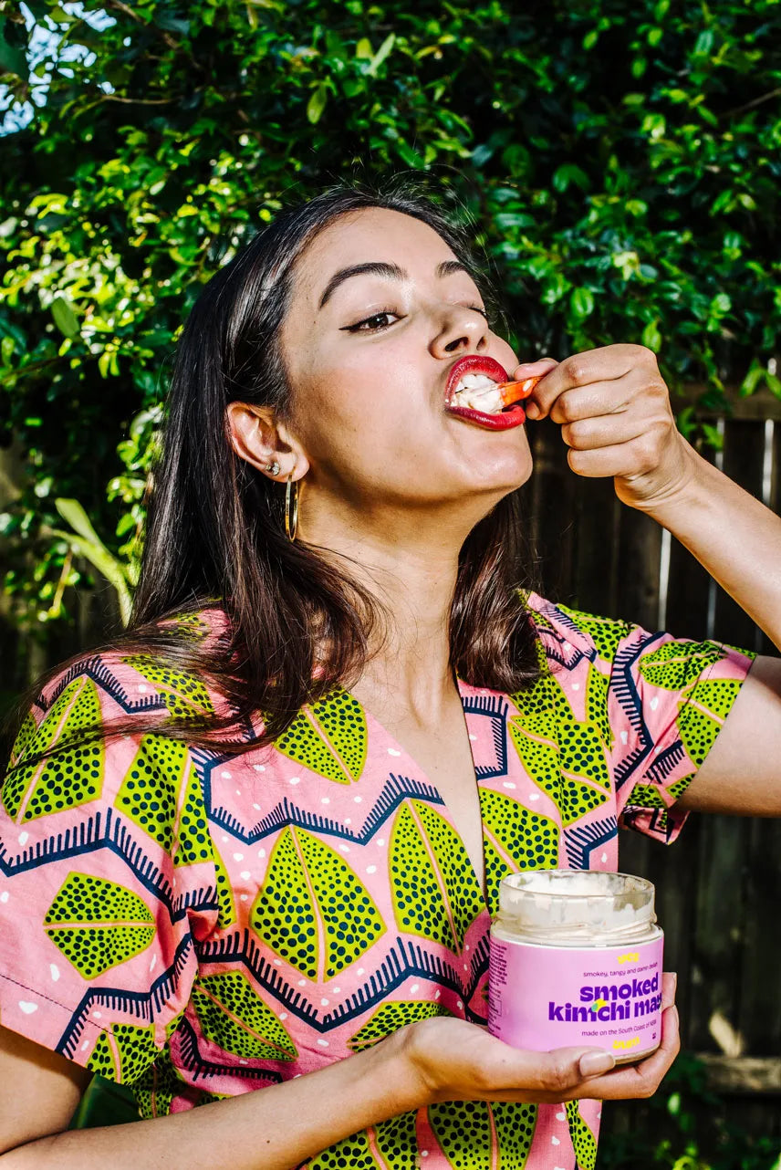Woman eating from a container labeled 'smoked kimchi' outdoors with greenery in the background