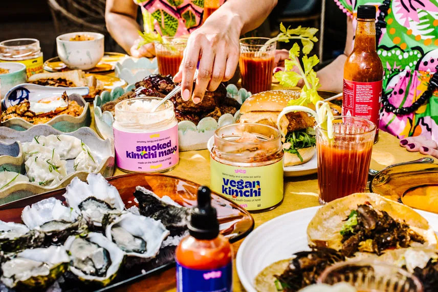 Table with various food items, drinks, and condiments with people partially visible.