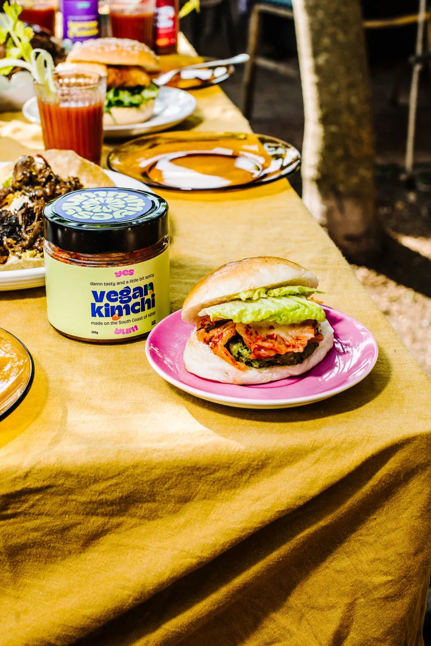 Vegan burger on a pink plate with a jar of 'vegan kimchi' on a yellow tablecloth.